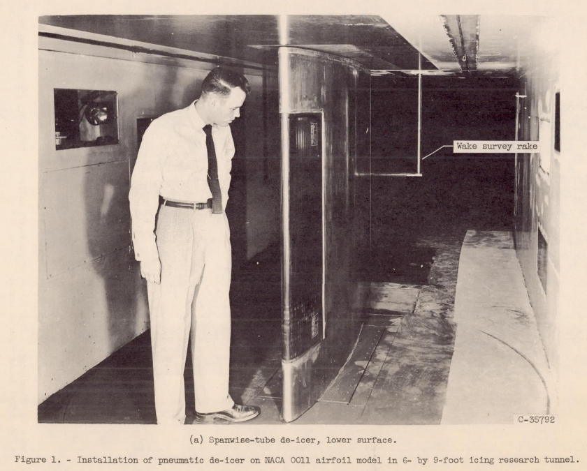A test airfoil mounted on a vertical axis in an icing wind tunnel, with a pneumatic de-icer covering part of the leading edge. A man stands next to it, viewing the leading edge. The caption reads:"Figure 1. Installation of pneumatic de-icer on the NACA 0011 airfoil model in 6 by 9 foot icing research tunnel. (a) Spawise-tube de-icier, lower surface. "